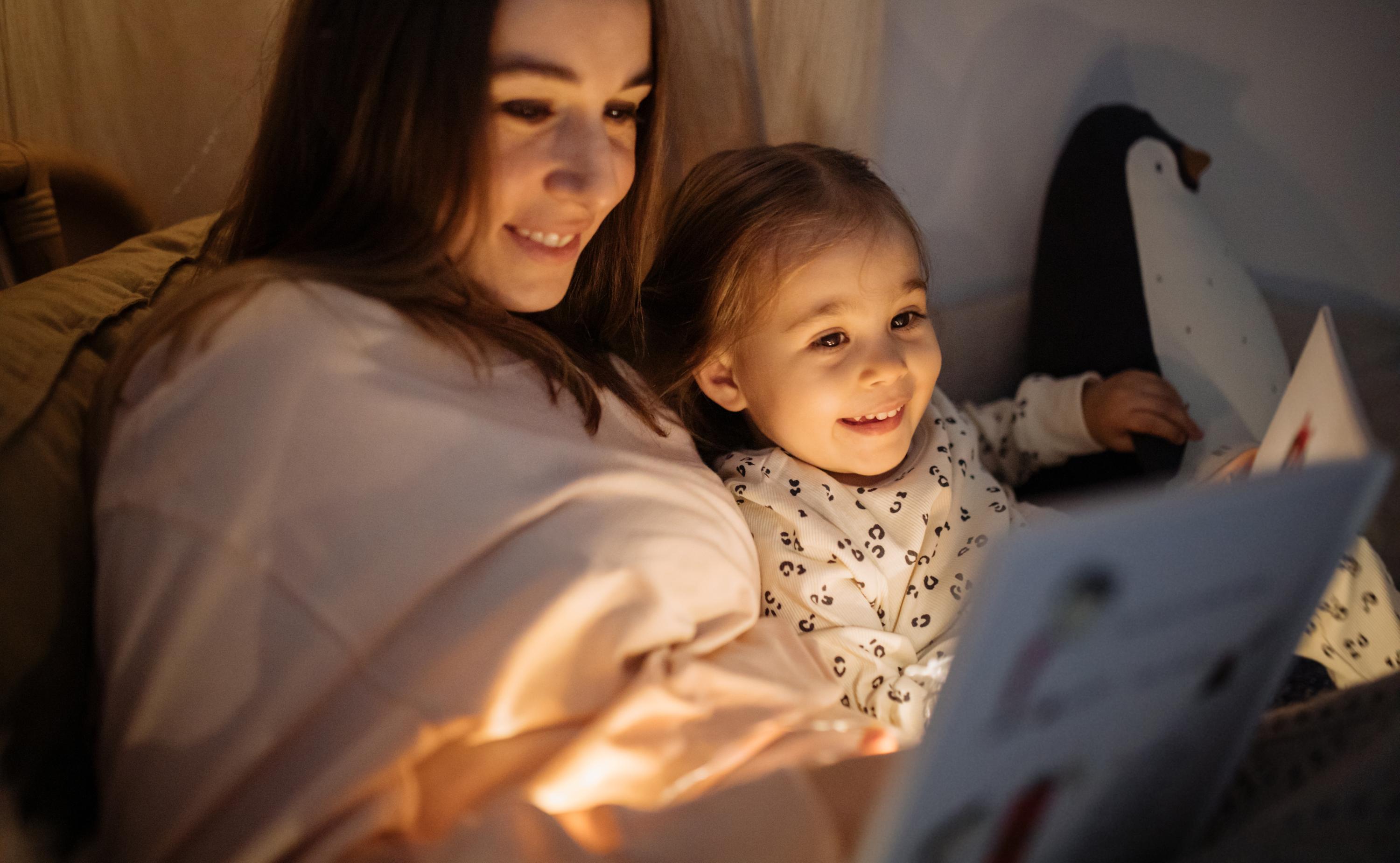 iStock-1296760408.jpg A woman lounging in a warmly dim lit space, reading to a child nestled in her arms.