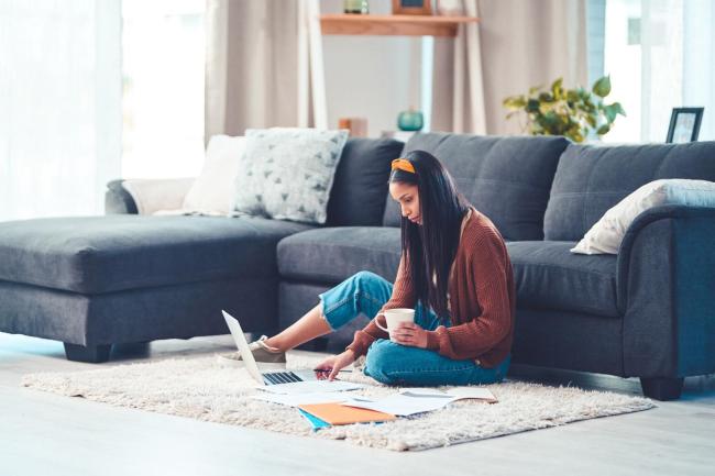 Woman in burnt orange-red sweater sitting on living room rug doing paperwork by her laptop