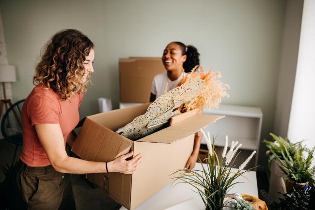 Two women roommates smiling in their new apartment as one holds a box of apartment decorations. 