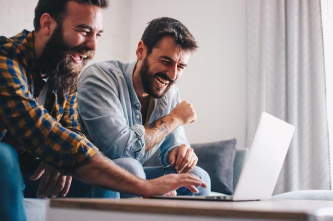 Two male roommates sitting in a living room and laughing as they look at a laptop. 