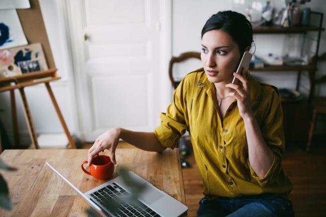 A young woman with her hair pulled back wearing a yellow shirt talks on the phone, while sitting at a table.