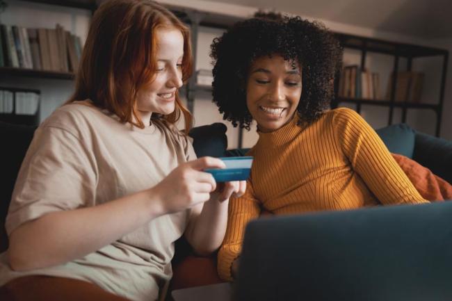 One woman holds a credit card up as another woman smiles as they makes a purchase on a laptop together. 