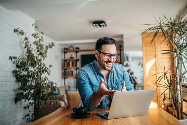 A cheerful man having a video call on laptop at a table in a white room with many green plants.