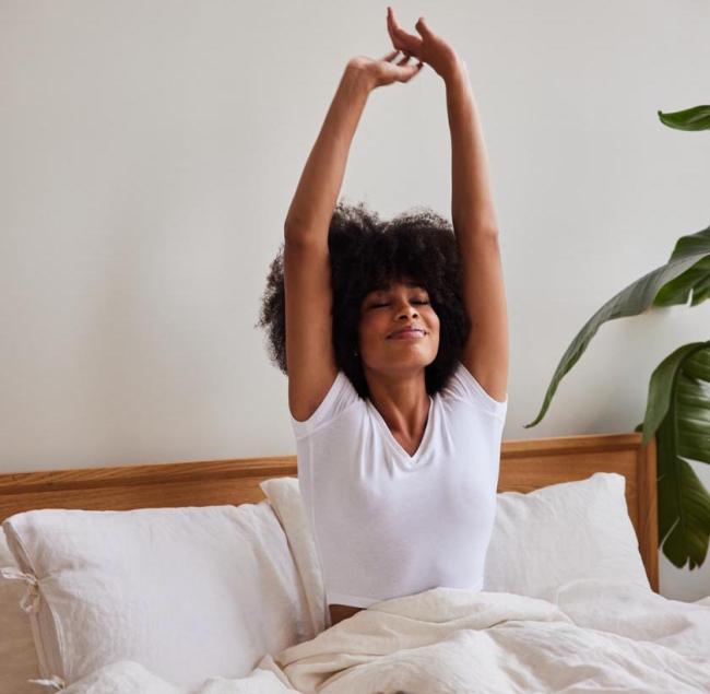 Young woman in a white shirt sitting up in bed stretching with both arms up in the air.