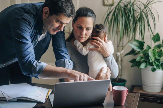Two young parents working from home, the man points at a laptop screen while the woman holds a baby. 