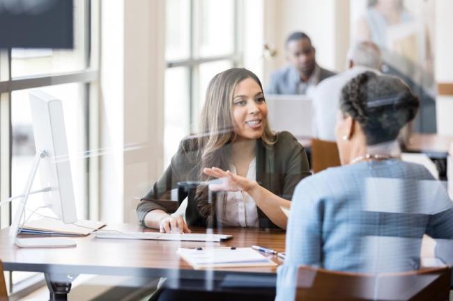 A banker dressed in a black blazer discusses banking services with a customer in an office.