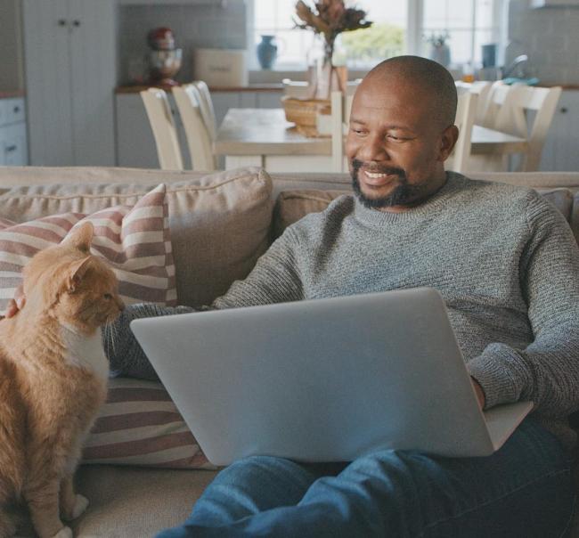 A man sitting on a couch smiling while he pets a cat and works on his laptop.
