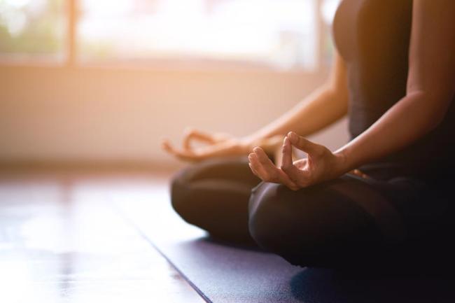 A woman sits on a yoga mat cross-legged with her hands in a zen pose resting on both knees. 