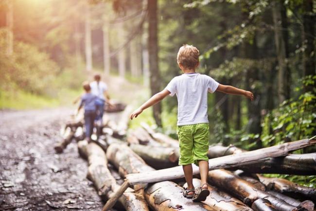 A young boy in green shorts and a white shirt walks carefully over a log outdoors with his arms held out for balance.