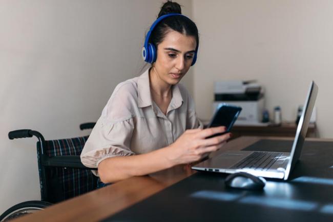 A young woman wearing headphones sits in a wheelchair at a table in front of a laptop holding her phone.