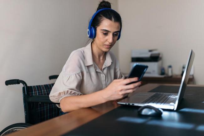 A young woman wearing headphones sits in a wheelchair at a table in front of a laptop holding her phone. 