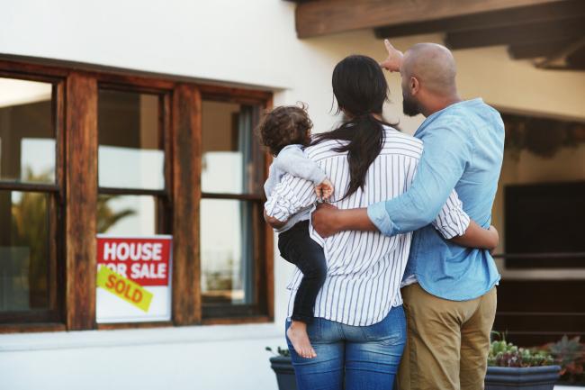 a couple with a small child, their backs turned to the camera and facing a home with a "for sale" sign.