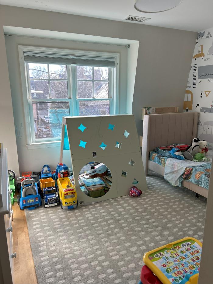 a bedroom with a large window and various playroom toys. Bedroom has off white walls and hardwood flooring.