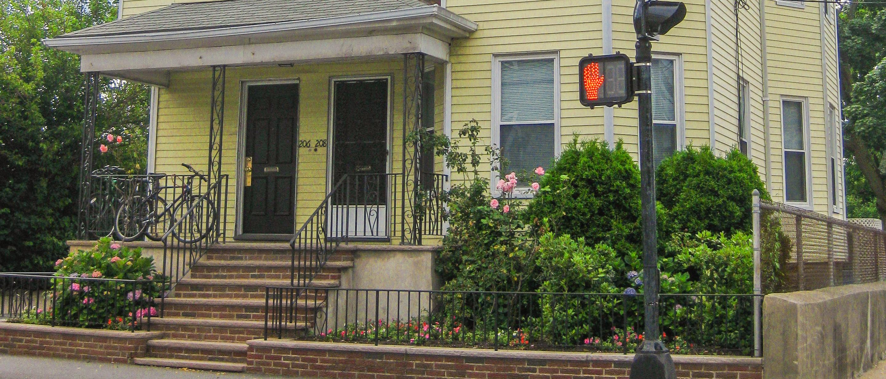 A yellow multifamily building located by a crosswalk. There is a small porch and landscaping surrounding the property.