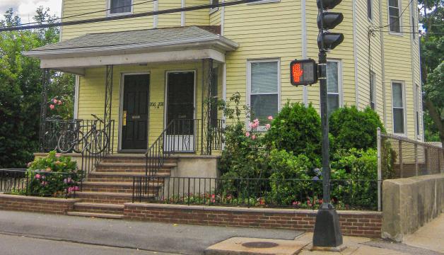 A yellow multifamily building located by a crosswalk. There is a small porch and landscaping surrounding the property.